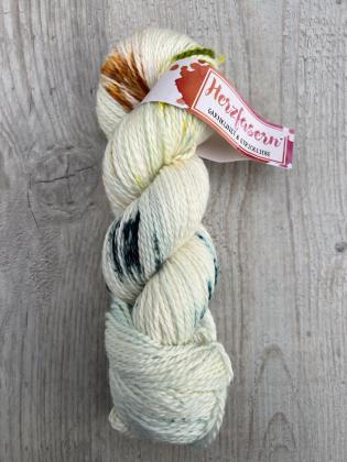 Ein Strang handgefärbtes Garn der Herzfaser No. 18, Grundfarbe naturweiß mit grünen, braunen und dunklen Sprenkeln. Liegt dekorativ auf hellem Holz.
